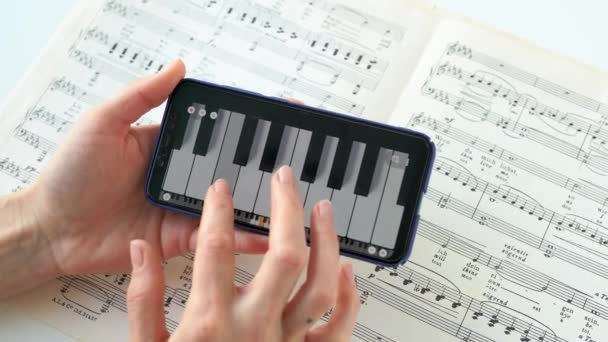 年轻女子正在练习用智能手机把手指按在键盘上弹钢琴 女孩的手弹奏着纸片音乐实践中撒谎笔记中的曲调 手机上的乐器 — 图库视频影像