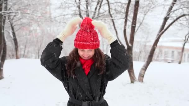 穿着白色针织手套的年轻漂亮的女人从戴着华丽帽子的红帽子上抖落雪花 看着相机笑了 在外面的雪地里 一个快乐的女孩笑着看着雪花 — 图库视频影像