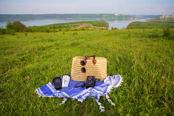 A very beautiful view of the lake. A straw picnic basket stands on a blue blanket on green grass along with glasses, a camera and coffee cups. Recreation in the open air