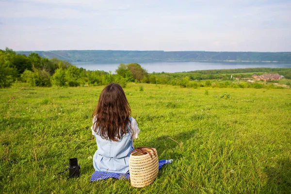 A very beautiful view of the lake, a girl is sitting on a blanket.. A straw picnic basket is standing nearby on the green grass along with an old camera. Recreation in the fresh air