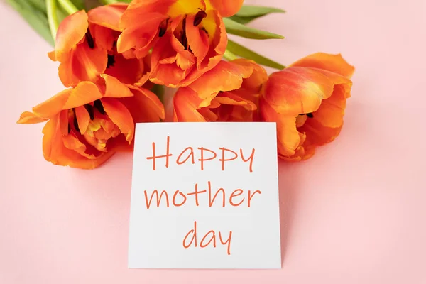 A beautiful bouquet of orange peony tulips along with a white card in the middle with the words happy mother day on a pink background
