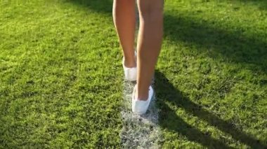 Beyaz spor ayakkabılı beyaz bir kız bir futbol sahasının beyaz çizgisi boyunca yürür. Çimenlerde yürüyen güzel bir kadının arka plan portresi. HD 25FPS 'te yavaş çekim video görüntüsü