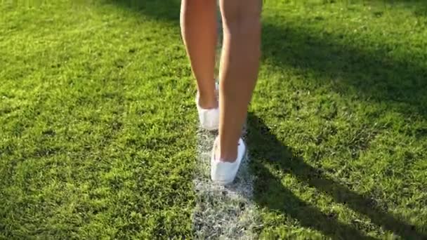 一个穿着白运动鞋的高加索女孩沿着足球场的白线走着 一个美丽的女人脚在草地上行走的背景肖像 25Fps慢镜头 — 图库视频影像