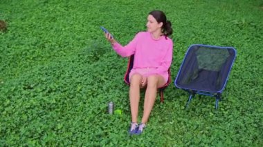 Doğada güneşlenen genç bir kadın. Termosun yanındaki sandalyede oturan mutlu kız selfie ya da görüntülü konuşma için akıllı telefon kullanıyor. Bir kadın yaz tatilinde açık hava yaşam tarzını sever.