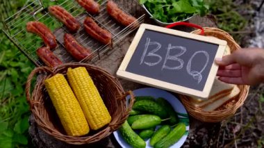 Izgara ızgaralı ağaç kütüğü, ızgara sosis, sebze, mısır, ekmek ve salatalık manzaralı yaz partisi. Kız eli karatahtaya yazılı barbekü tahtası koyuyor. 4k 25FPS içinde video