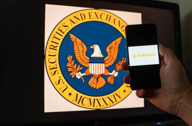 Kyiv, Ukrayna - 06 Haziran 2023: Adam şifreli bir akıllı telefonu SEC logosu olan monitörün arka planına karşı ekranda Binance 'ı takas ediyor. S.E.C. BNB 'de para aklama yoluyla Binance' a dava açılmış.