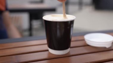 Caddedeki bir kafede yavaş ve hızlı karıştırılmış şeker. Elinizde ahşap bir kaşık var ve kapuçinoyu beyaz süt köpüğüyle karıştırın. Çekim için şehir kahvesi