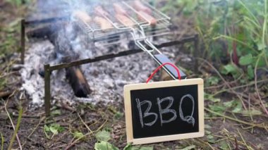 Baharat ve otlarla ızgarada pişen lezzetli sosisler. Tebeşirle Barbekü yazılı küçük karatahtayla kamp yapmak. Yaz mevsiminde arka bahçede piknik. Video görüntüleri 4k 25FPS