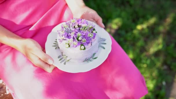 美しい女性の手は 自然のプレート上に白い紫色のケーキをねじれています ピンクのドレスを着た誕生日の女の子屋外 ケーキを持っている女性は 鐘や花を飾り 彼の指でクリームを試してみてください — ストック動画