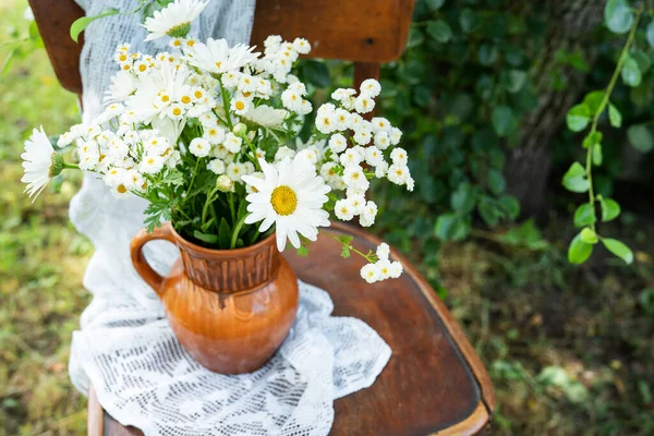 Summer atmosphere, simple home decor in the countryside. Beautiful white daisies on a wooden old chair along with a lace napkin against the backdrop of a green garden