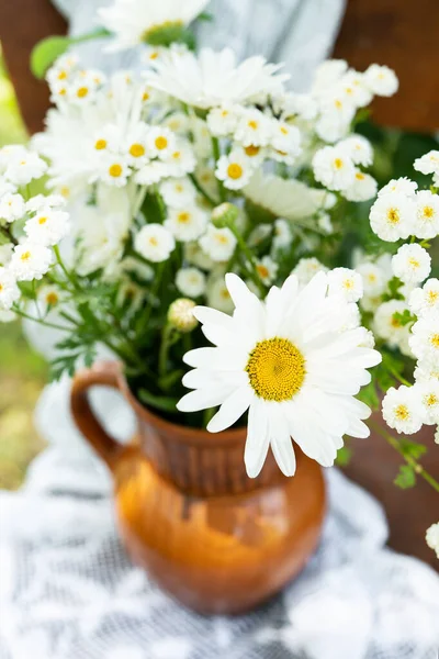 Beautiful chamomile flowers on a wooden old chair along with a lace doily on a green garden background. Summer atmosphere, simple home decor in the countryside. Vertical photo