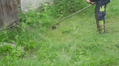 Koruyucu üniformalı bahçıvan kırsal alanda ot biçiyor. İşçi, petrol çim biçme makinesi ya da elektrikli tırpan ile uzun çimleri biçiyor ve bitki örtüsünün küçük parçaları farklı yönlere dağılıyor.