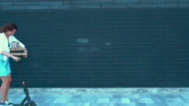 Genç bayan kurye, e-scooterıyla paket teslim ediyor. Elektrikli scooter kullanan mutlu bir kadın, yerleşim bölgesinde sokakta bir sürü karton kutu taşıyor. Teslimat ve çevre dostu modern yaşam tarzı