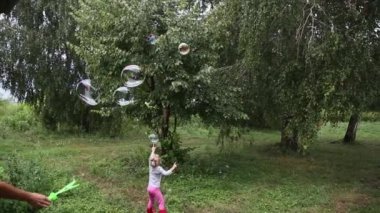 Dışarıda sabun köpüğüyle oynayan mutlu küçük kız. Neşeli bebek gülüyor, zıplıyor, annesinin ağaçların arasında çimenlikte üflediği kabarcıkları yakalıyor. Komik çocuk doğada balon üfleyiciyle yakalıyor.