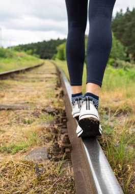 Kız raylar boyunca yürüyor, dikiz aynasından. Kızın bacakları rayların üzerinde, yakın plan. Raylarda yürümenin tehlikesi