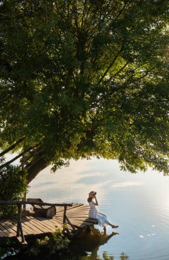 Şapkalı ve uzun elbiseli genç bir kız gölün kıyısındaki bir rıhtımda oturur ve doğan güneşe bakar. Seyahat ve açık hava eğlencesi kavramı.