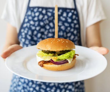 Önlüklü bir kadın şef, geleneksel bir hamburgeri çedar peyniri ve salatayla nasıl servis ettiğini gösteriyor. Yakın plan.