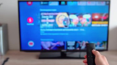 Televizyonun uzaktan kumandasını tutan ve oturma odasındaki odaksız akıllı televizyondaki kanalları çeviren kadın elinin yakın çekimi. İnternet kanalında sörf yapan bir kadın. 4K 'da ev sineması