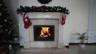Noel arifesinde yanan yakacak odunları olan beyaz mermer şömine. Yeni yıl süslemeleri, yanıp sönen çelenkler ve hediye olarak çoraplarla çevrili. Sıcak ateşin önünde evde rahatlatıcı bir sahne. 4K