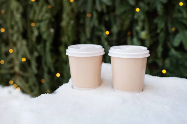 2つのプラスチックカップは クリスマスツリーの緑の壁とガーランドからのボケの背景に雪の上に立っています コーヒー 新鮮な空気の中で散歩する — ストック写真