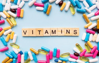 Çeşitli renkli haplar ve kapsüller parlak mavi bir arka plana saçılmış. VITAMINS kelimesi ahşap kiremitler kullanılarak yazılır.