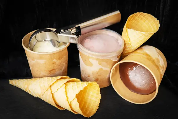 This image shows three cups of ice cream in different flavors: vanilla, strawberry and chocolate. On one of the cups lies a metal ice cream scoop, three empty waffle cones stacked against each other