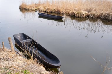 Durgun bir gölde iki siyah tekne. Kuru otlarla çevrili. Bulutlu gökyüzünü yansıtıyor.