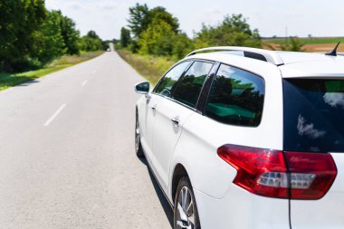 Güneşli bir yolda park edilmiş beyaz bir araba yeşillikler ve açık gökyüzü arasında bir yolculuğa hazır. Sahne huzur ve macera dolu.