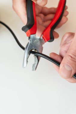 Beyaz arka plandaki siyah kabloyu kesmek için kırmızı ve siyah pense kullanan ellerin yakın çekimi.