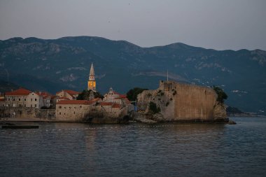 Karanlık çöktüğünde sahil kasabasında tarihi binalar ve arka planda dağlara karşı kurulan deniz kenarı kalesi. Budva, Karadağ