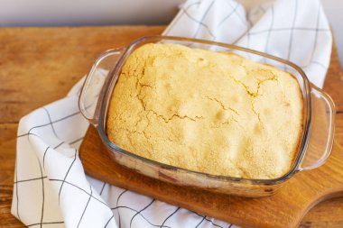 Hafif çatlamış bir kapağı olan altın kahverengimsi bir tatlı, ahşap bir tahtanın üzerinde cam bir pişirme kabında oturur..