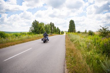 Motosikletli bir motorcu kırsal alanlar arasında yol boyunca gidiyor. Yaz, güneşli gün, güzel gökyüzü