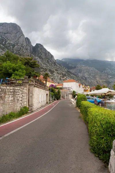Bulutlu bir gökyüzünün altında dramatik dağlara yuva yapmış pitoresk bir sahil kasabası, üzerinde bisiklet yolu olan asfalt bir yola sahip..