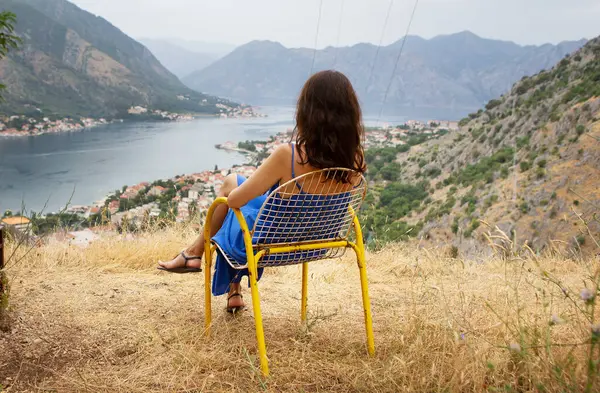 Mavi elbiseli bir kadın sarı bir sandalyede dinleniyor, dağlarla çevrili sakin bir körfezin nefes kesici manzarasının tadını çıkarıyor..