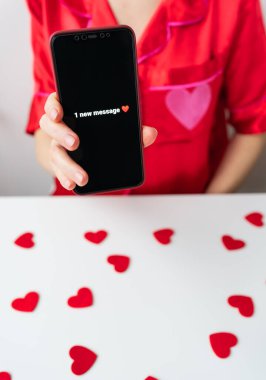 Bir kız elinde bir akıllı telefon tutuyor ve içinde 1 yeni mesaj var yazıyor. Telefona odaklanmış durumda ve kız beyaz arka planda kırmızı bir gömlek giyiyor.