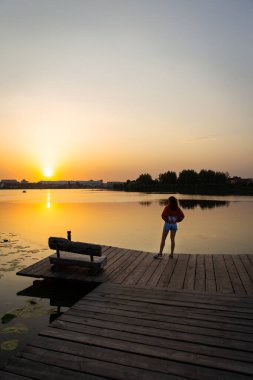 Gün batımında manzaralı bir göl manzarası Tahta bir iskelede duran bir kadın, Tahta bir bank ve sakin su üzerinde altın yansımalar.