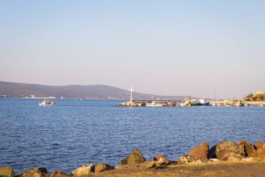 Küçük bir limanı, tekneleri ve berrak bir gökyüzünün altında uzak bir dağ sırası olan sakin bir deniz manzarası..