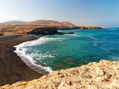 Fuerteventura adasındaki Ajuy kasabasında siyah volkanik kumlu bir plaj bu resimde gösterilmektedir. Engebeli kıyı şeridi berrak turkuaz sularla buluşurken, arka planda çorak tepeler yükseliyor..