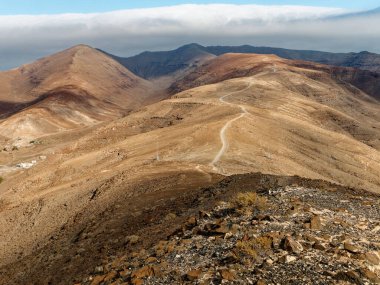 Fuerteventura 'nın en yüksek dağı Pico de la Zarza' nın zirvesine giden yol kurak ve kayalık arazide esiyor. Dalgalanan manzara, arka planda bulutlu bir ufuk olan uzak tepeleri ortaya çıkarıyor..