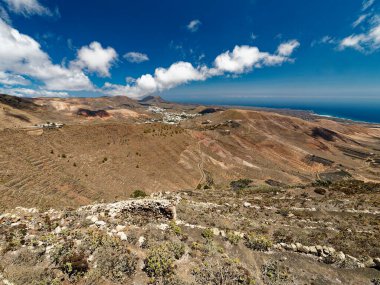 Lanzarote volkanik manzarası, katmanlı tepeleri ve seyrek bitki örtüsüyle çorak ve engebeli bir araziye sahiptir. Uzak okyanus, bulutlarla dağılmış berrak mavi gökyüzünün altındaki kuru toprakla çelişiyor..