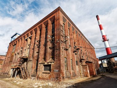 Görüntü, yıpranmış dokuları olan büyük, kırmızı tuğlalı bir endüstriyel binayı gösteriyor. Kenarına bir merdiven bağlanmış. Arka planda, kırmızı ve beyaz bir baca parçalı bulutlu bir gökyüzüne doğru yükselir..