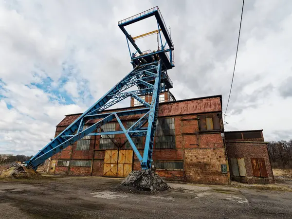 Görüntü, Pszw 'daki eski bir sanayi yapısı olan kömür madeninin bacasını gösteriyor. Mavi metal çerçeve bulutlu bir gökyüzüne karşı yükseliyor, paslı ve yıpranmış detaylarla tuğla bir binaya bağlanıyor. Sahne terk edilmiş gibi..