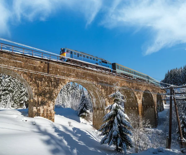 Stone viaduct (arch bridge) on railway through mountain snowy fir forest and locomotive  with a passenger train. Snow drifts  on wayside and hoarfrost on trees and electric line wires.