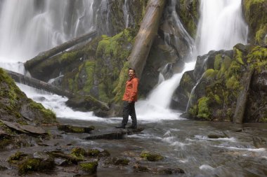 Oregon ormanında büyük ve güzel bir şelalenin önünde duran turuncu ceketli bir yürüyüşçü.