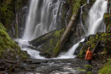 Oregon ormanında büyük ve güzel bir şelalenin önünde duran turuncu ceketli bir yürüyüşçü.