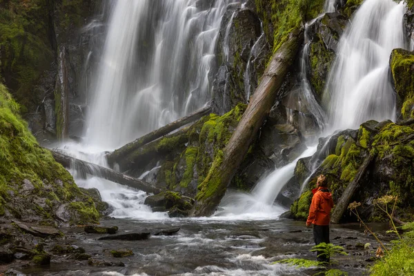 Oregon ormanında büyük ve güzel bir şelalenin önünde duran turuncu ceketli bir yürüyüşçü.