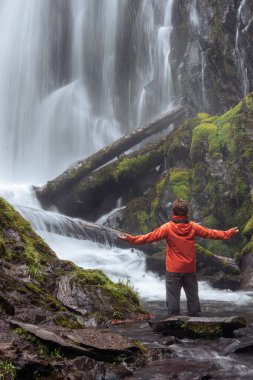 Oregon ormanında büyük ve güzel bir şelalenin önünde duran turuncu ceketli bir yürüyüşçü.