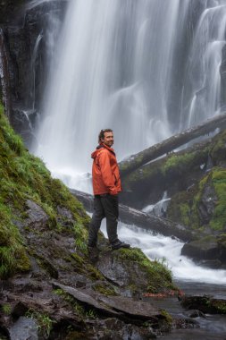 Oregon ormanında büyük ve güzel bir şelalenin önünde duran turuncu ceketli bir yürüyüşçü.