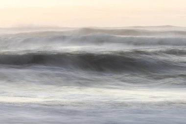 Görüntü, sahile çarpan dalganın bulanık bir görüntüsü. Su beyaz ve gökyüzü pembe. Huzurlu ve rahatlatıcı bir atmosfer yaratıyor. Dalgalar kıyıya çarpıyor.