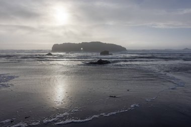 Okyanus sakin ve gökyüzü bulutlu. Güneş bulutların arasında parlıyor. Kumsal kayalık ve su sakin.